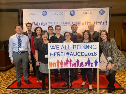 A group of faculty, staff students and alumni together in front of a sign that says “We all belong here! # AUCD 2018”