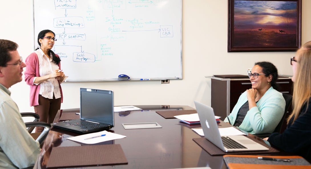 A researcher standing with a whiteboard with writing behind her and 3 individuals at a table in front of her with laptops, papers and pens